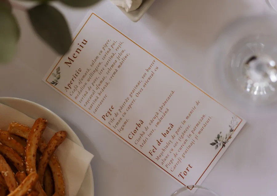 Meniu de nuntă personalizat, cu chenar auriu și frunze, pe masa festivă lângă un platou cu covrigei